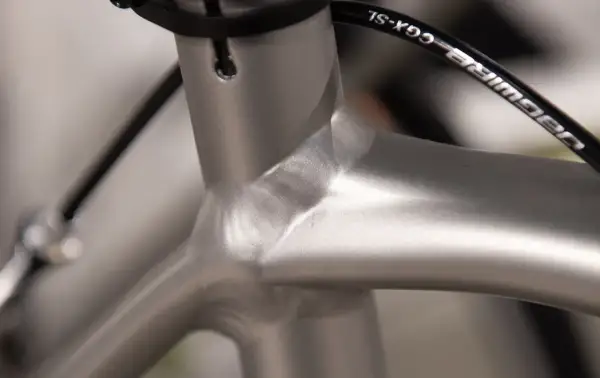 Close-up van de balhoofdbuis van een racefiets. Het frame is van aluminium, met een gladde, zilverkleurige afwerking. De kabels van de remmen lopen langs het frame.