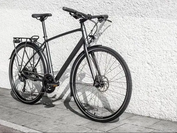 Grijze, moderne custom fiets met bagagedrager en schijfremmen, leunend tegen een witte, gestructureerde muur.