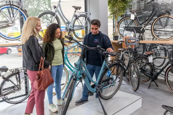 Een verkoper toont een lichtblauwe Gazelle bedrijfsfiets aan twee vrouwen in een fietsenwinkel.
