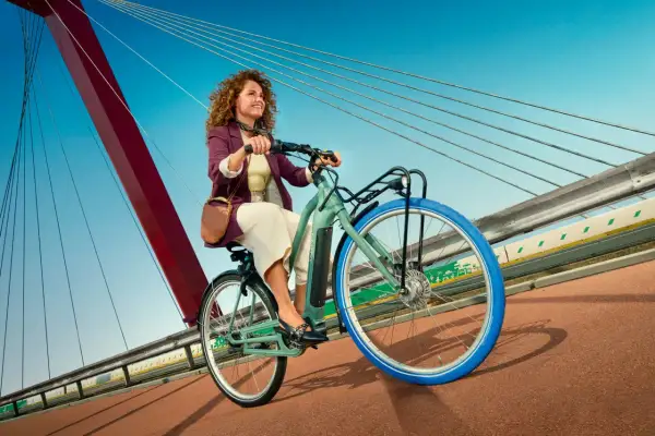 Vrolijke vrouw met krullend haar rijdt op een groene Swapfiets over een brug met blauwe lucht.