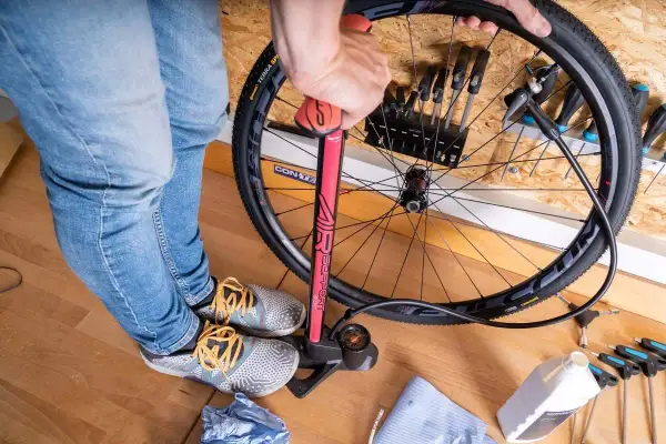 Een persoon pompt een tubeless band op met een rode fietspomp. Gereedschap en een flesje vloeistof liggen klaar.